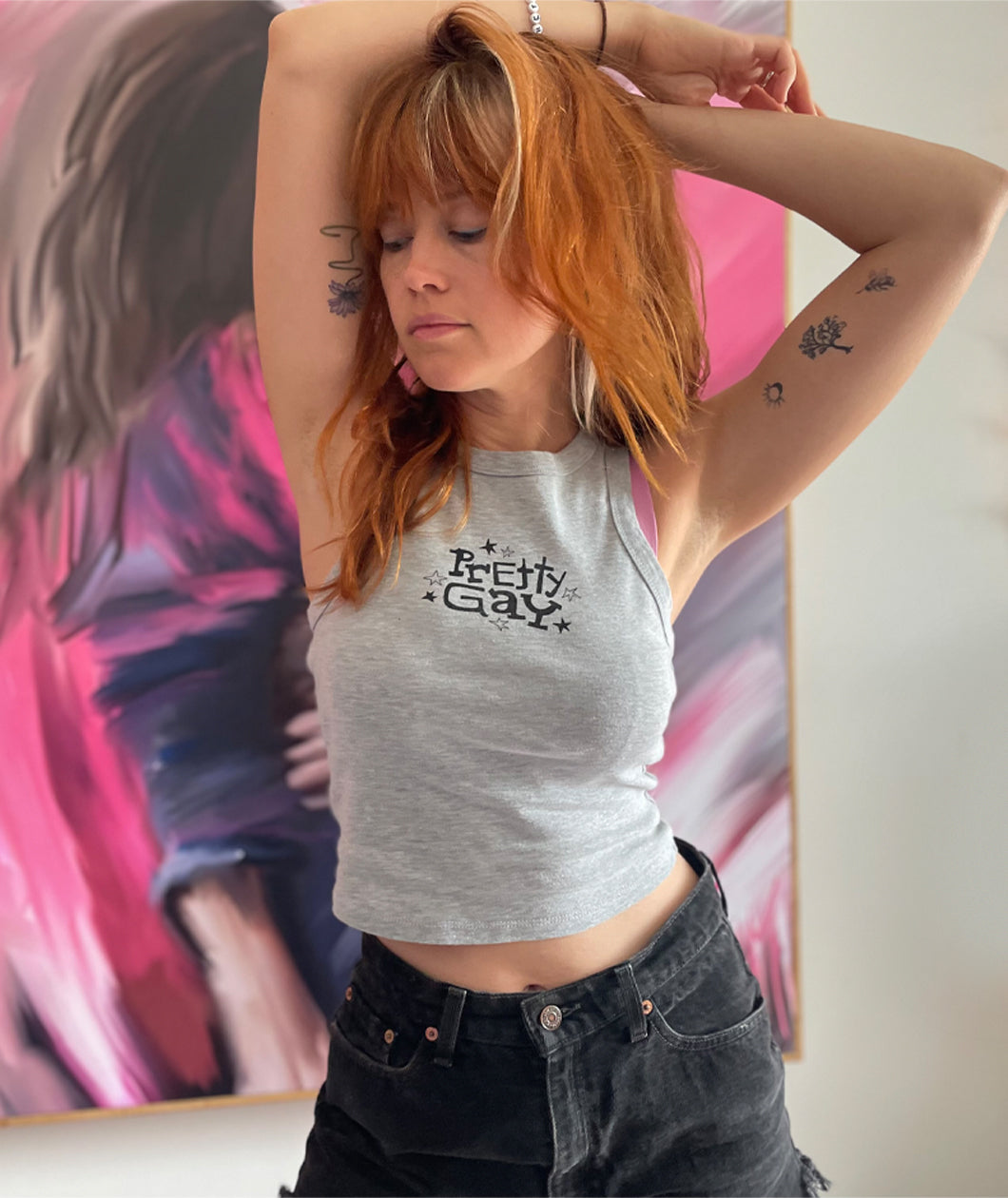A photo of Catherine McCafferty posing, wearing a cropped gray tank top. The tank top reads "Pretty Gay" and has little star illustrations around it. From Pretty Gay.