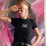 A photo of Catherine McCafferty posing, wearing a black t-shirt. The shirt reads "Pretty Gay" and has little star illustrations around it. From Pretty Gay.