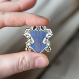 Gold enamel pin held between someone's thumb and forefinger to show size. Pin is about 1.25" tall and depicts a powder blue corset surrounded by gold, white, and pink daises with green leaves and a light blue background.