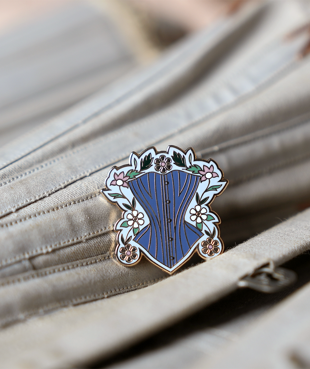 Pin is about 1.25" tall and depicts a powder blue corset surrounded by gold, white, and pink daises with green leaves and a light blue background. Pin is resting on a grey corset