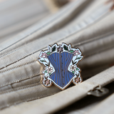 Pin is about 1.25" tall and depicts a powder blue corset surrounded by gold, white, and pink daises with green leaves and a light blue background. Pin is resting on a grey corset