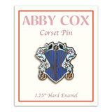 Pin is about 1.25" tall and depicts a powder blue corset surrounded by gold, white, and pink daises with green leaves and a light blue background. Pin is on a paper card backer with Abby Cox written on the top with text "Corset Pin" beneath and then "1.25" Hard Enamel" written below the pin.