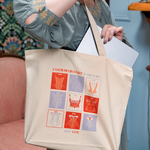 Woman in a blue dress holding a canvas tote with red and light purple square design on the front. Text says FASHION HISTORY IS HISTORY and then under the design says ABBY COX. She is placing a laptop into the bag which seems to take up about half of the space.