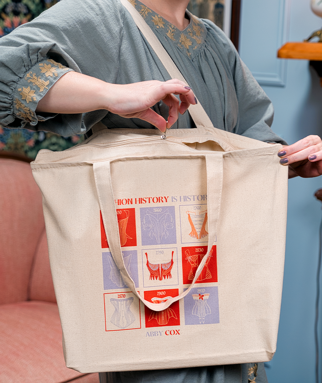 A woman zipping the canvas tote with red and light purple square design on the front. Text says FASHION HISTORY IS HISTORY and then under the design says ABBY COX.