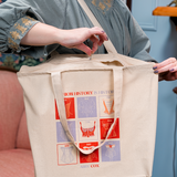 A woman zipping the canvas tote with red and light purple square design on the front. Text says FASHION HISTORY IS HISTORY and then under the design says ABBY COX.