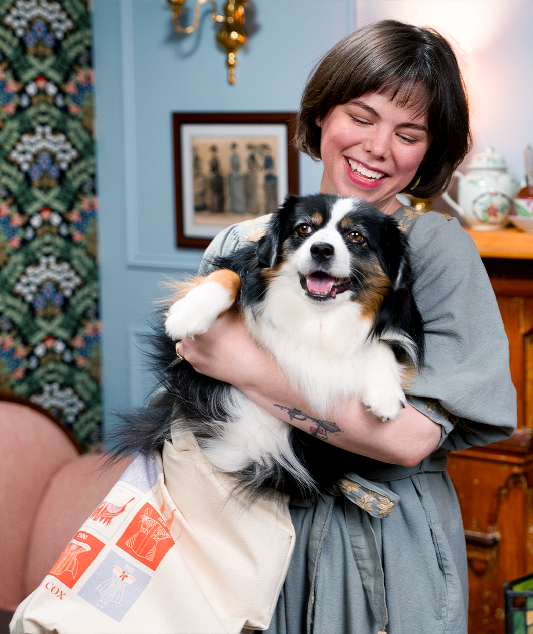 A woman with a short black bob haircut is smiling holding a white and black fluffy puppy that is half in a canvas tote. The dog is also smiling.