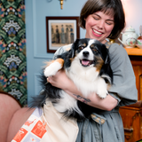 A woman with a short black bob haircut is smiling holding a white and black fluffy puppy that is half in a canvas tote. The dog is also smiling.