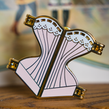 Gold hard enamel pin shaped like a corset filled with pale pink enamel and a white enamel top edge to look like lace with gold detailing. The pin is split down the middle with golden hinges on each of the four corners to show it can open.