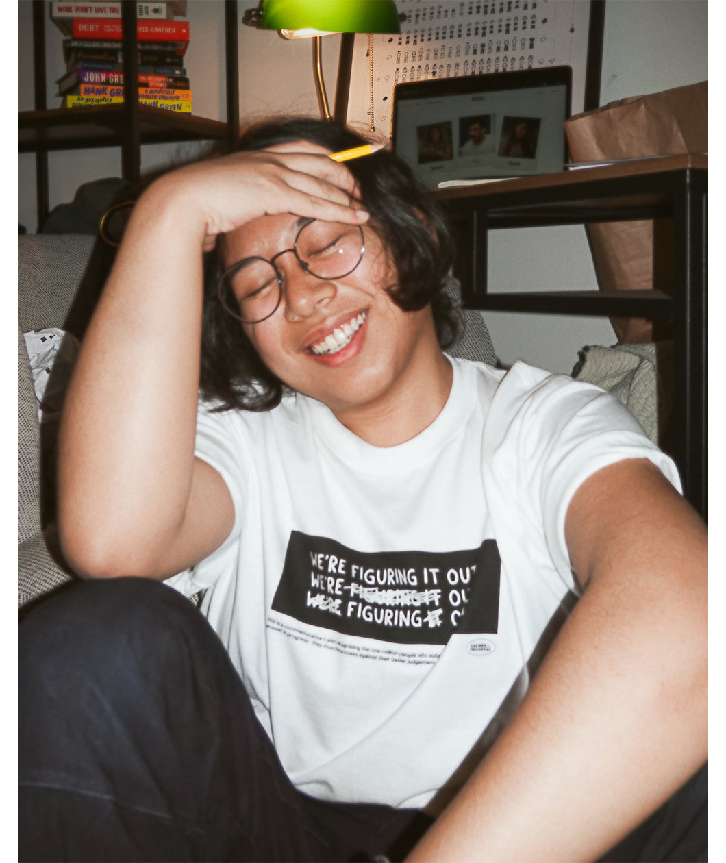 A photo of person sitting on the ground, smiling with their eyes closed wearing a white t-shirt with a black rectangle in the center with the words "We're figuring it out" written three times in a row with the second line has "figuring" crossed out and the third line has "we're" and "it" crossed out. There is additional small text below this. From Answer in Progress.