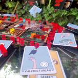 Close up of the role and character cards showing the Valedictorian in this game is the Secret Stabber. The background has the game box and the room cards with stab tokens scattered.