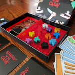 Open game box showing the bright red interior, red stab tokens, 10 colorful meeples, and the deck of cards with the front of the box in the background. Stabbin in the Cabin box is glossy black on the outside. There are some cards in the box and some cards fanned out around the box.