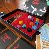 Open game box showing the bright red interior, red stab tokens, 10 colorful meeples, and the deck of cards with the front of the box in the background. Stabbin in the Cabin box is glossy black on the outside. There are some cards in the box and some cards fanned out around the box.