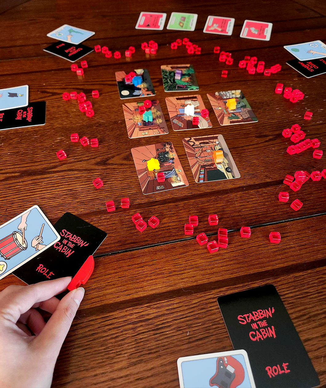 Stabbin in the Cabin board game set up on a wooden table ready to play. The room cards are arranged in a grid and the role and character cards surround the room cards with the red translucent stab tokens in between. A hand holds the knife token and multiple meeples lay dead on the room cards.