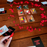 Stabbin in the Cabin board game set up on a wooden table ready to play. The room cards are arranged in a grid and the role and character cards surround the room cards with the red translucent stab tokens in between. A hand holds the knife token and multiple meeples lay dead on the room cards.