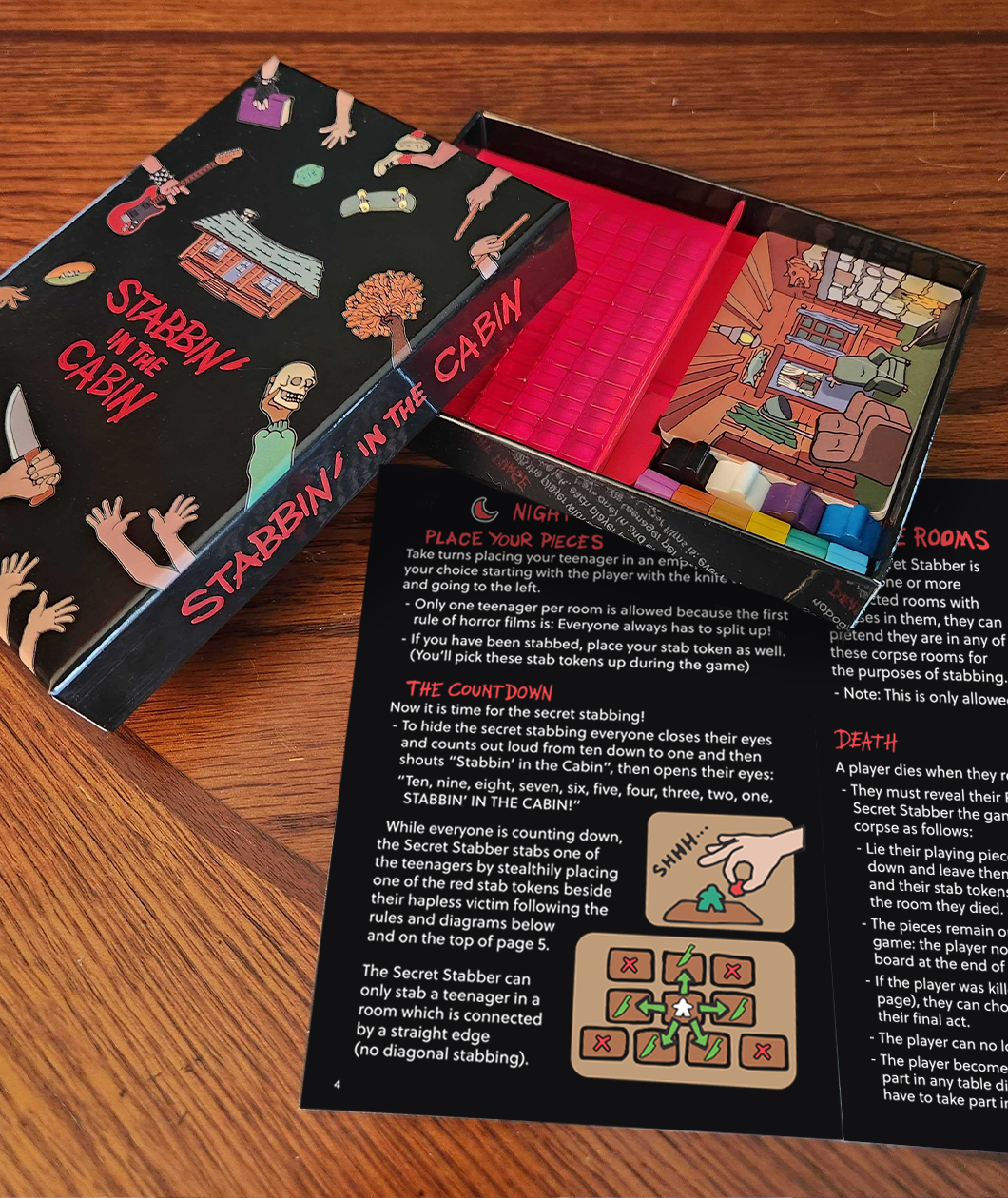 Stabbin in the Cabin Board game box and instructions booklet on a wooden table. Bo is open so you can see the 100 red translucent cubes, 10 meeples, and deck of cards. Outside of the box is a glossy black with the character printed on it and Stabbin in the Cabin in bright red in a drippy bloodlike font. Inside of the box is bright red. Instructions booklet is black with white and red text.
