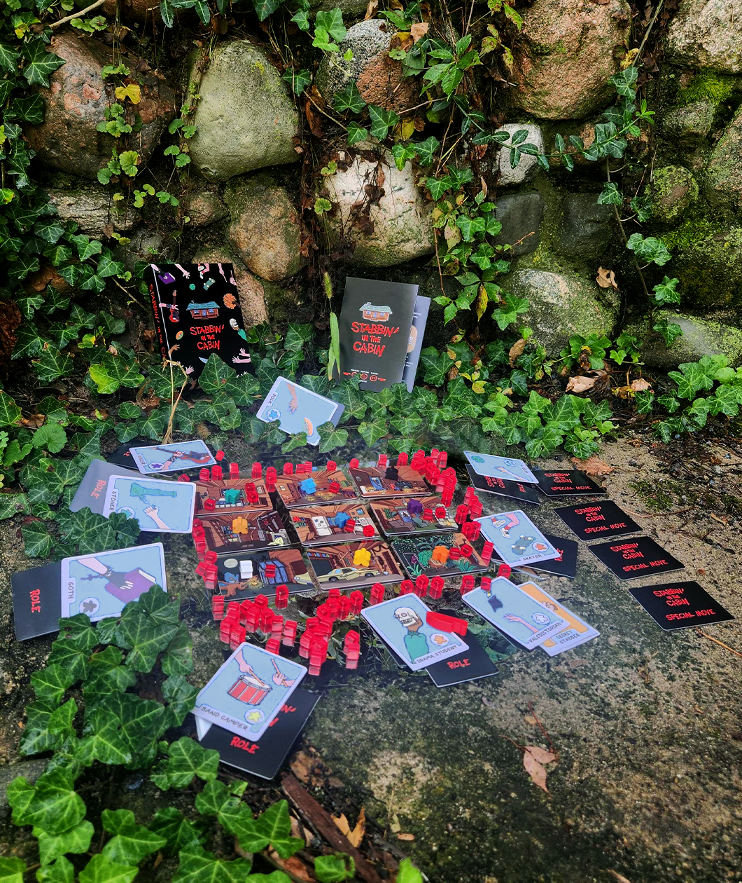 Stabbin in the Cabin board game set up outside on ivy and stone ready to play. The room cards are arranged in a grid and the role and character cards surround the room cards with the red translucent stab tokens in between.