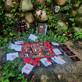 Stabbin in the Cabin board game set up outside on ivy and stone ready to play. The room cards are arranged in a grid and the role and character cards surround the room cards with the red translucent stab tokens in between.