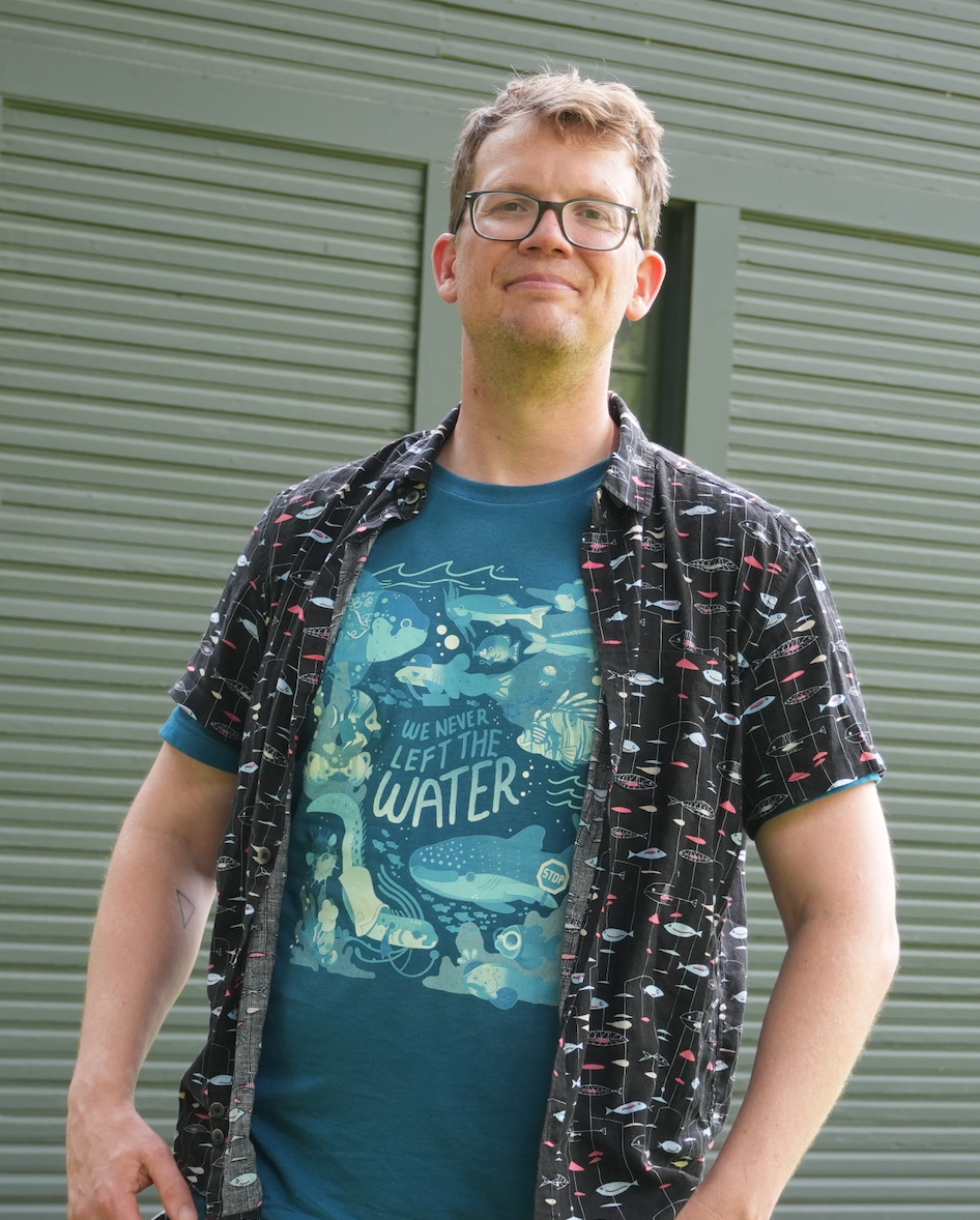 A white man with glasses who calls himself Hank Green, wearing a deep teal tshirt that reads, "WE NEVER LEFT THE WATER" with fish illustrations. He is wearing a black button up shirt open over the tshirt that has a red and blue pattern of fish on it.