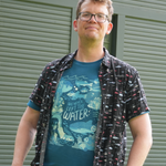 A white man with glasses who calls himself Hank Green, wearing a deep teal tshirt that reads, "WE NEVER LEFT THE WATER" with fish illustrations. He is wearing a black button up shirt open over the tshirt that has a red and blue pattern of fish on it.