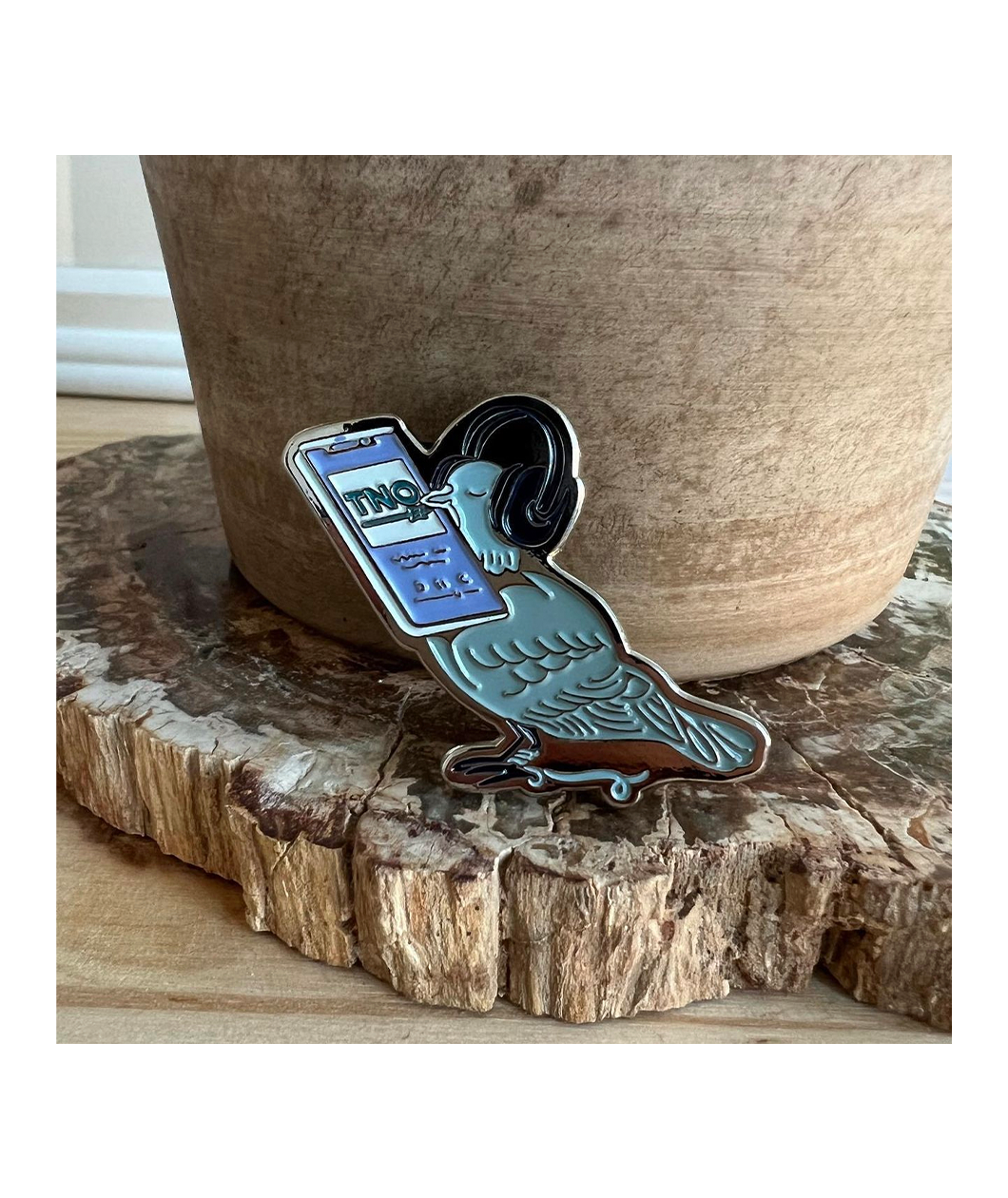 An enamel pin shaped like a blue bird wearing headphones, holding a cellphone that is playing TNO.