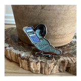 An enamel pin shaped like a blue bird wearing headphones, holding a cellphone that is playing TNO.