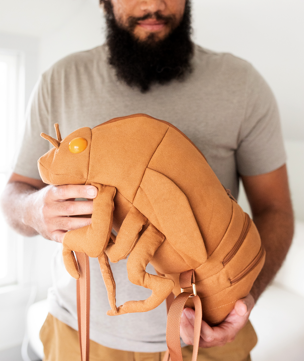Man holding canvas backpack in the shape of a cicada shell complete with brown straps that attach the front legs to the thorax and egg yolk shaped protruding eyes. 