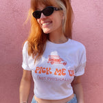 Catherine McCafferty is wearing a white crop top and sunglasses in front of a pink wall. The crop top has large text that reads, "Pick me up (not physically)" in orange on the front. Above the text is an illustration of a pick up truck with text on the side that reads, "Pretty Gay". She is smiling and has her thumbs in the loops of her jeans. From Pretty Gay.