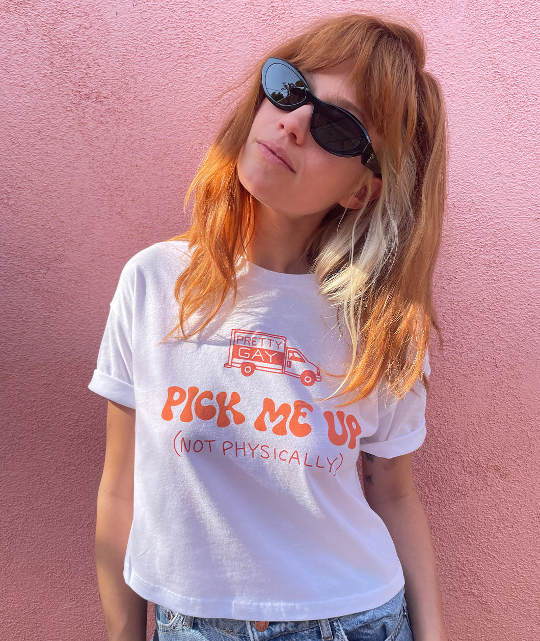 Catherine McCafferty is wearing a white crop top and sunglasses in front of a pink wall. The crop top has large text that reads, "Pick me up (not physically)" in orange on the front. Above the text is an illustration of a pick up truck with text on the side that reads, "Pretty Gay". From Pretty Gay.