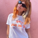 Catherine McCafferty is wearing a white crop top and sunglasses in front of a pink wall. The crop top has large text that reads, "Pick me up (not physically)" in orange on the front. Above the text is an illustration of a pick up truck with text on the side that reads, "Pretty Gay". From Pretty Gay.