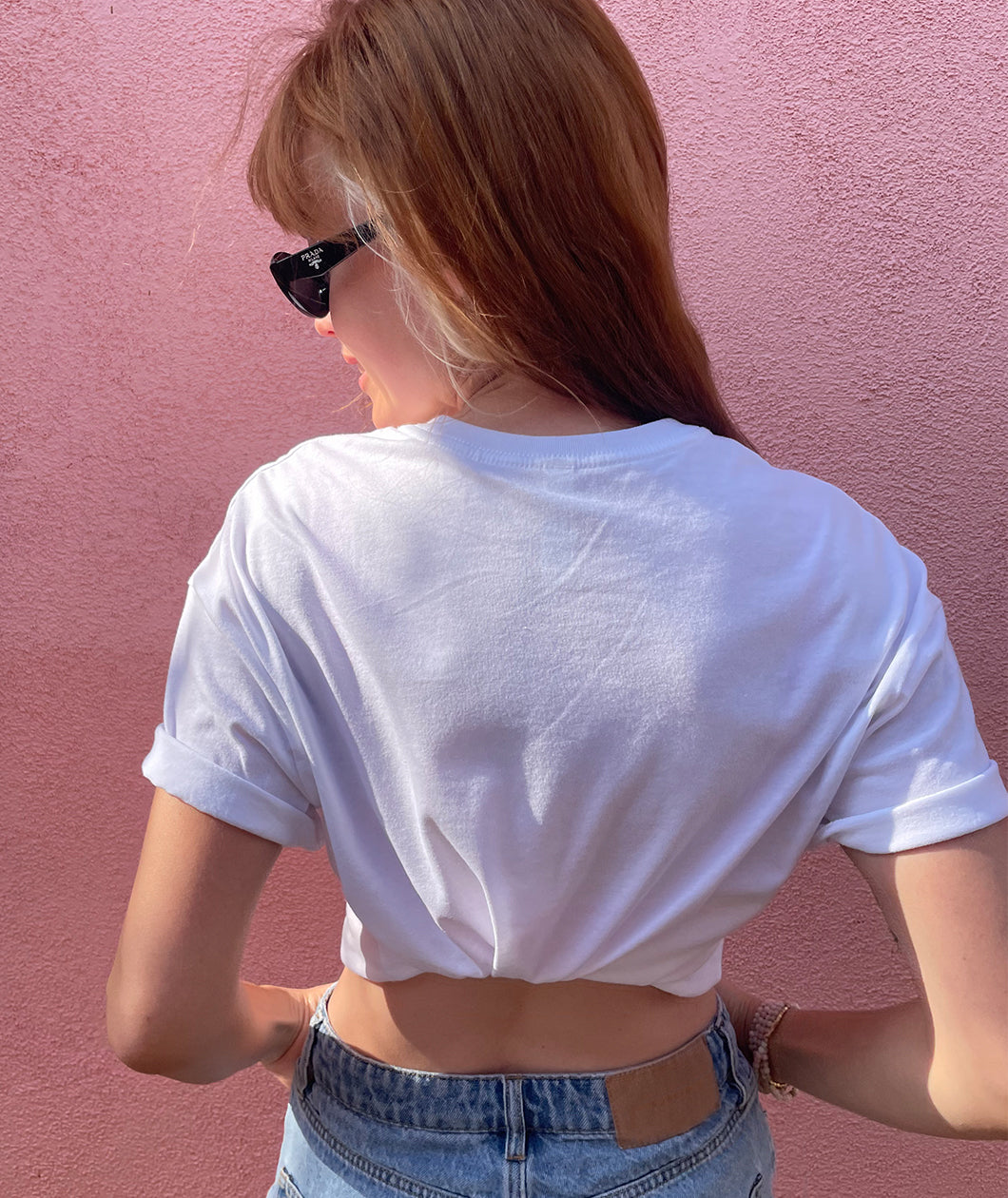 Catherine McCafferty is wearing a white crop top and sunglasses in front of a pink wall. She is turned facing away to show how the crop top can be gathered in the back. From Pretty Gay.