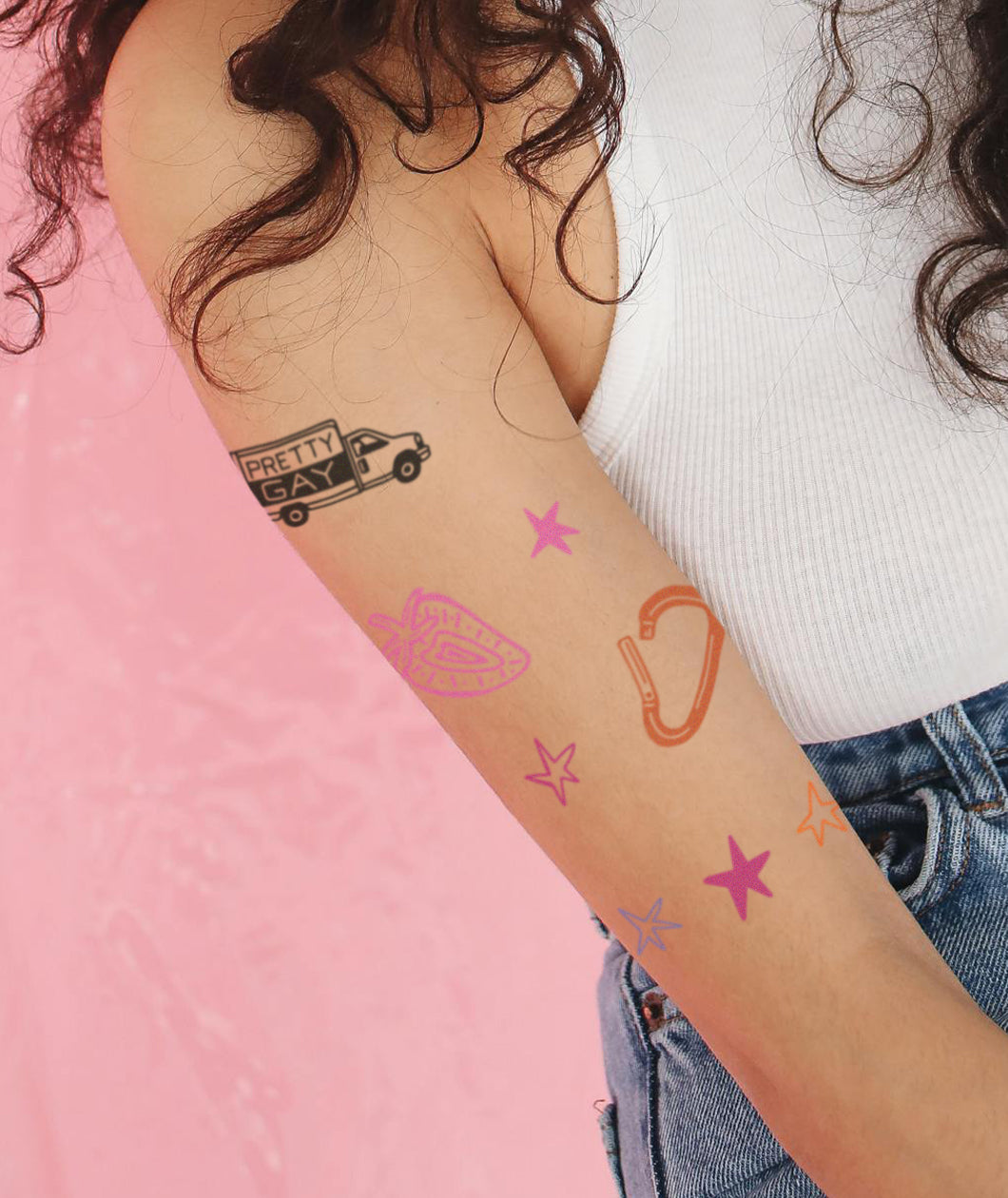 A closeup of a woman's upper arm that has several temporary tattoos applied. The tattoos include a truck that says "Pretty Gay", a strawberry, a carabiner, and several stars. From Pretty Gay.
