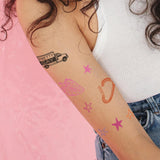 A closeup of a woman's upper arm that has several temporary tattoos applied. The tattoos include a truck that says "Pretty Gay", a strawberry, a carabiner, and several stars. From Pretty Gay.