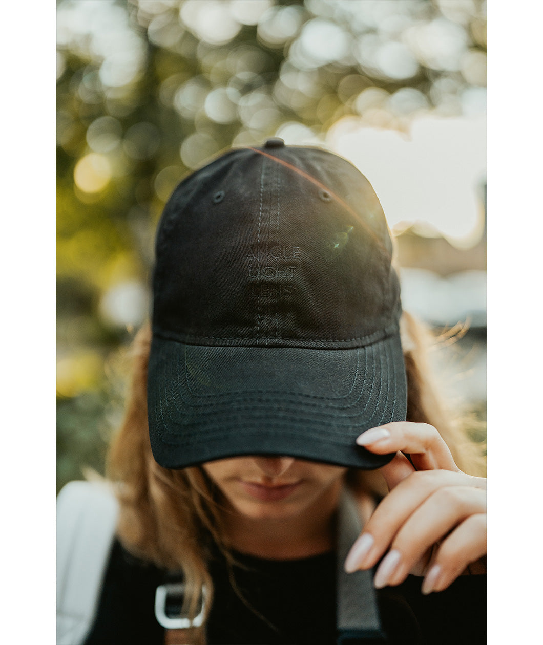 Lizzie Peirce wearing a hat with her head pointed down. The hat is black and is embroidered with "Angle Light Lens."