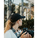 Lizzie Peirce wearing a hat, looking into the distance and holding a camera. The hat has a small white crescent embroidered on the back.