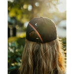 Lizzie Peirce wearing a hat, with her back to the camera. The hat has a small white crescent embroidered on the back.