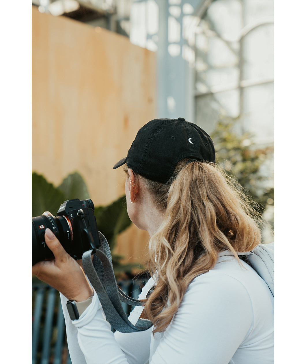 Lizzie Peirce wearing a hat, looking into the distance and holding a camera. The hat has a small white crescent embroidered on the back.