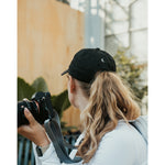 Lizzie Peirce wearing a hat, looking into the distance and holding a camera. The hat has a small white crescent embroidered on the back.