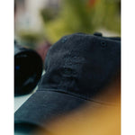 A close up of a black hat embroidered with "Angle Light Lens" sits on a table with a camera next to it.