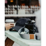 A black hat embroidered with "Angle Light Lens" sits on a table with a camera next to it.