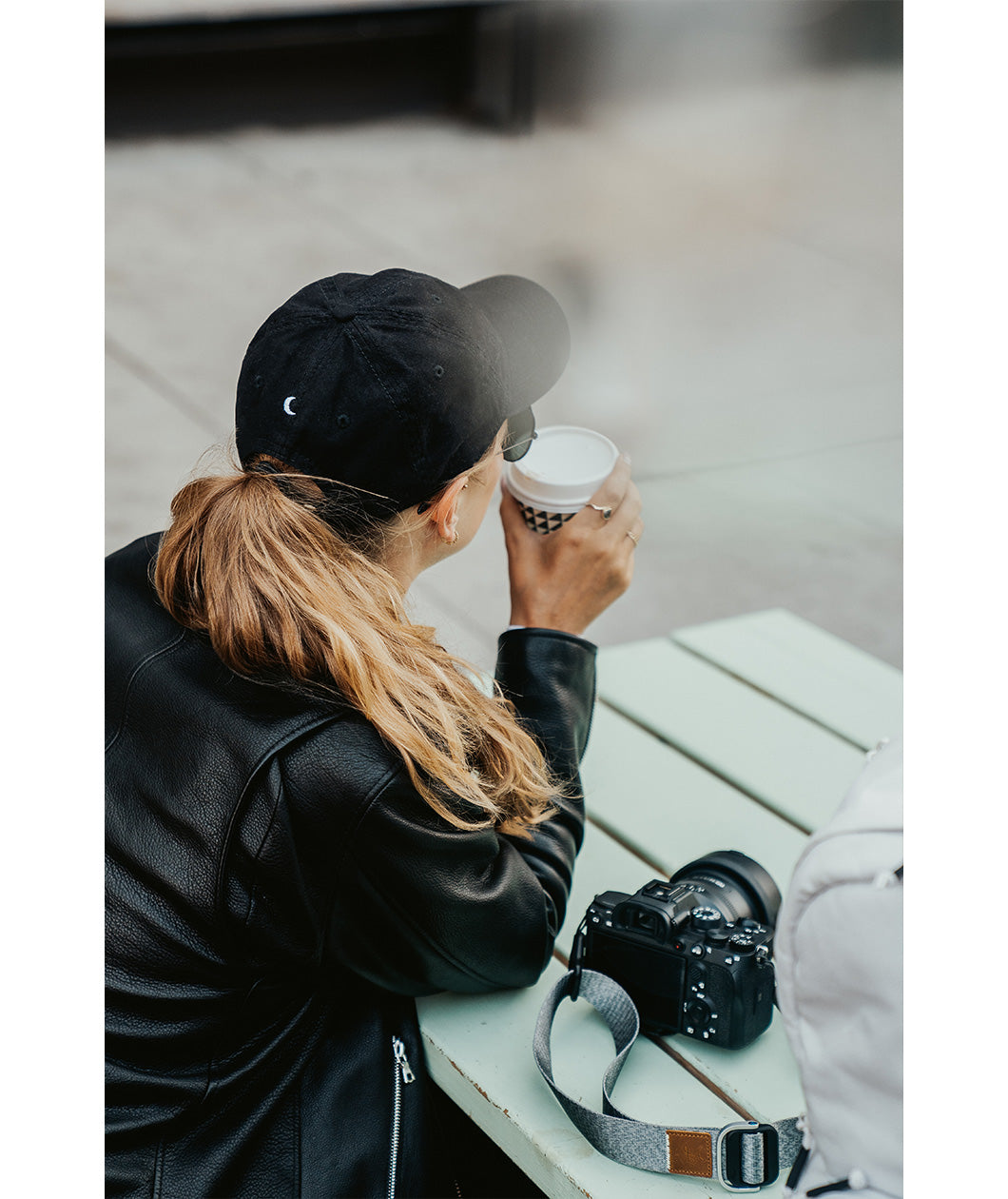 Lizzie Peirce wearing a hat, facing away from the camera, drinking coffee with a camera on the table next to her. The hat is black and is embroidered with a small white crescent moon on the back.