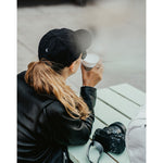 Lizzie Peirce wearing a hat, facing away from the camera, drinking coffee with a camera on the table next to her. The hat is black and is embroidered with a small white crescent moon on the back.