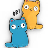 Yellow cartoon cat pin with a thin black outline. Two small paws at the bottom with a tail coming out at the bottom right. Tongue is hanging out and eyes are white with two black X’s in its eyes. Blue cartoon cat pin with black outline. Two small paws are at the bottom with a tail coming out of the bottom left. Two white eyes with black pupils looking off in opposite direction - from Physics Girl