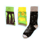 A set of three different socks. First: Brown sock folden in half with a green ankle and three leaves. Second: Light green sock folded in half with yellow ankle with a varying styles of yellow dandelions. Third: A black sock with tan ankle, heel, and toe dandelion multiple dandelion seeds floating away from dandelion - from This Star Won’t Go Out