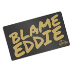 Black rectangular playmat with curved corners. “Blame Eddie” is in gold font resembling handwriting. “The commander’s quarters” is in the bottom right corner in gold sans serif font with a gold signature above it - from The Commander’s Quarters