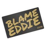Black rectangular playmat with curved corners. “Blame Eddie” is in gold font resembling handwriting. “The commander’s quarters” is in the bottom right corner in gold sans serif font - from The Commander’s Quarters