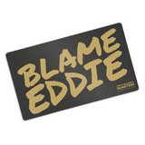 Black rectangular playmat with curved corners. “Blame Eddie” is in gold font resembling handwriting. “The commander’s quarters” is in the bottom right corner in gold sans serif font - from The Commander’s Quarters
