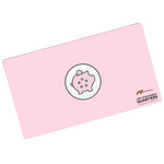 A pink rectangular playmat with curved corners. In the center is a white circle with a black outline. A piggy bank is in the center with a coin being inserted. On the pig are five circles of different colors. “The Commander’s Quarters” is in the bottom right in black sans serif font with a signature above - from The Commaner’s Quarters
