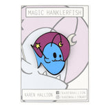 A magic Hanklerfish pin on a grey backing card. The pin is a purple circle with a blue hanklerfish inside holding a baton with a star. From Karen Hallion.