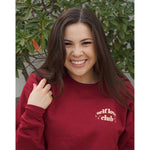 A woman modeling a red crewneck sweatshirt with “Self love” in white serif font outlined in orange arched above “club” in white serif font outline in orange. “Club” is surrounded by orange stars - from Sierra Schultzzie