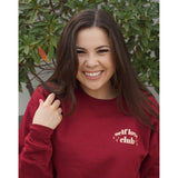 A woman modeling a red crewneck sweatshirt with “Self love” in white serif font outlined in orange arched above “club” in white serif font outline in orange. “Club” is surrounded by orange stars - from Sierra Schultzzie